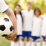 Several children standing together, excitedly surrounding a soccer ball in a sunny outdoor setting.