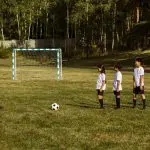 Several children standing together, excitedly surrounding a soccer ball in a sunny outdoor setting.
