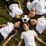 A family of children lying on the grass, joyfully playing with a soccer ball under a clear blue sky.
