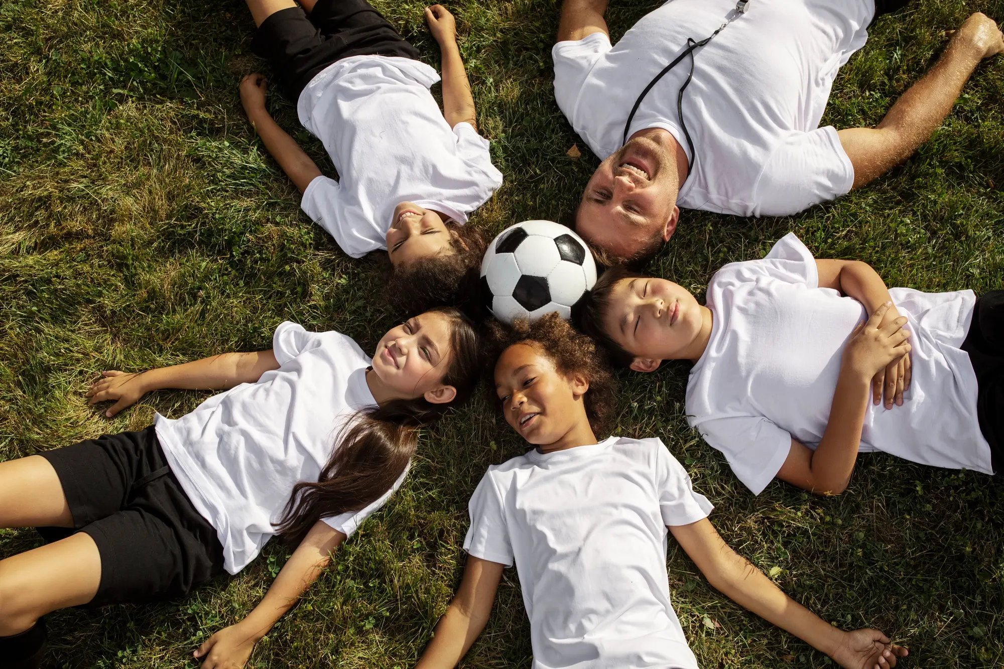 A family of children lying on the grass, joyfully playing with a soccer ball under a clear blue sky.