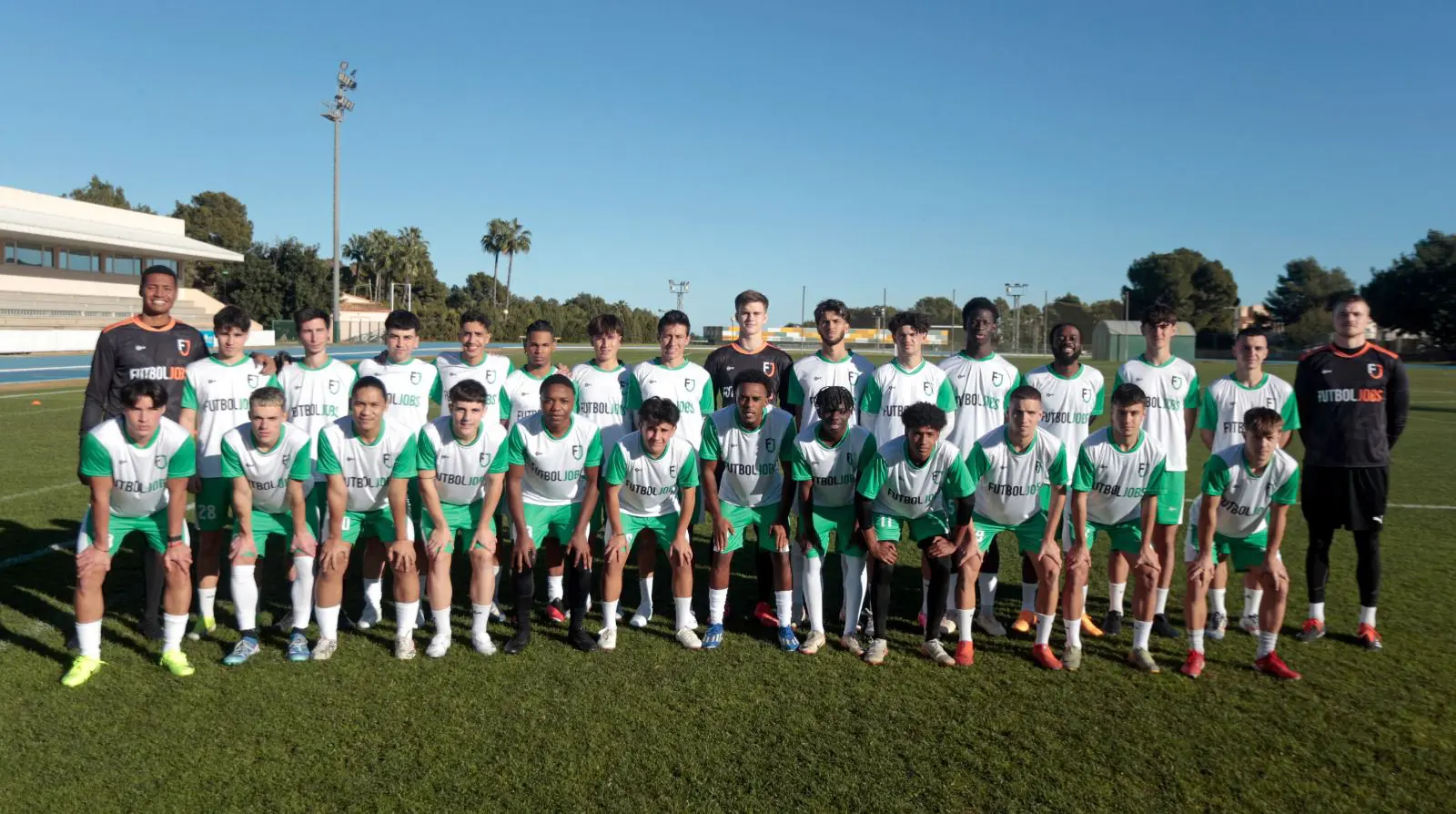 A soccer team stands together on the field, smiling for a group photo in their uniforms.