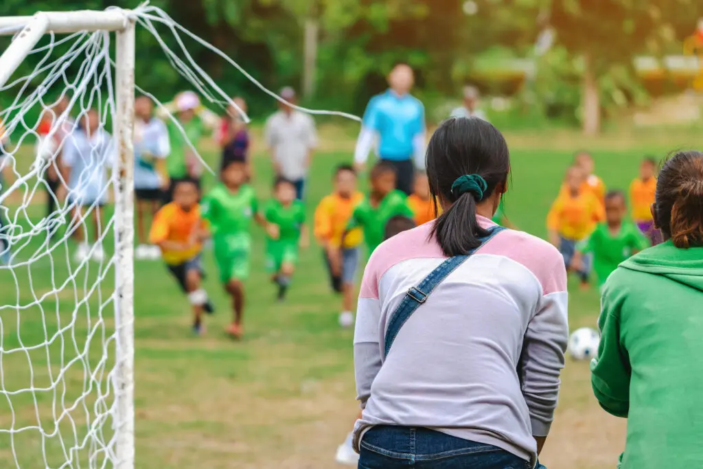 Back view of Moms watch and cheering their sons playing football in school tournament on sideline. Sport, outdoor active, Spectator watching soccer game. Parents care and encourage their children.