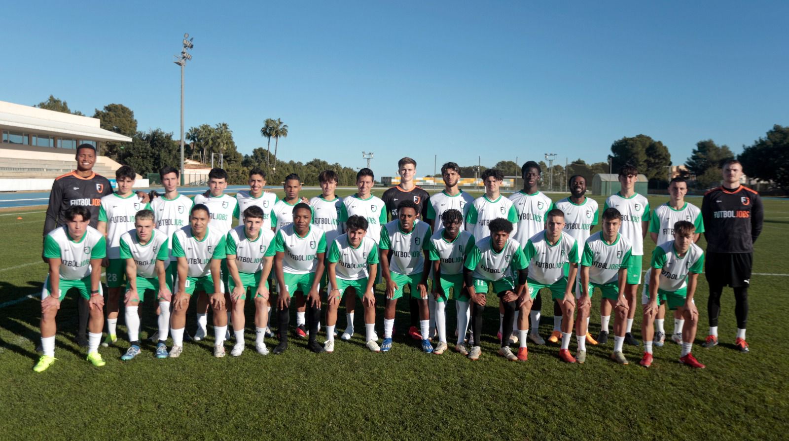 A soccer team stands together on the field, smiling for a group photo in their uniforms.