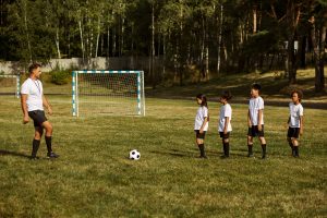 Several children standing together, excitedly surrounding a soccer ball in a sunny outdoor setting.