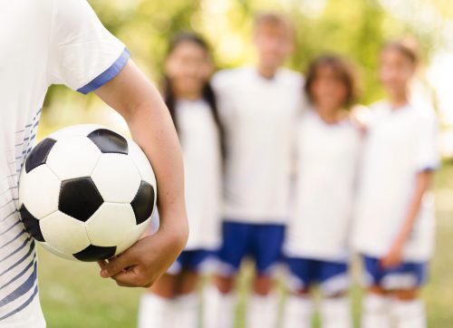 Several children standing together, excitedly surrounding a soccer ball in a sunny outdoor setting.
